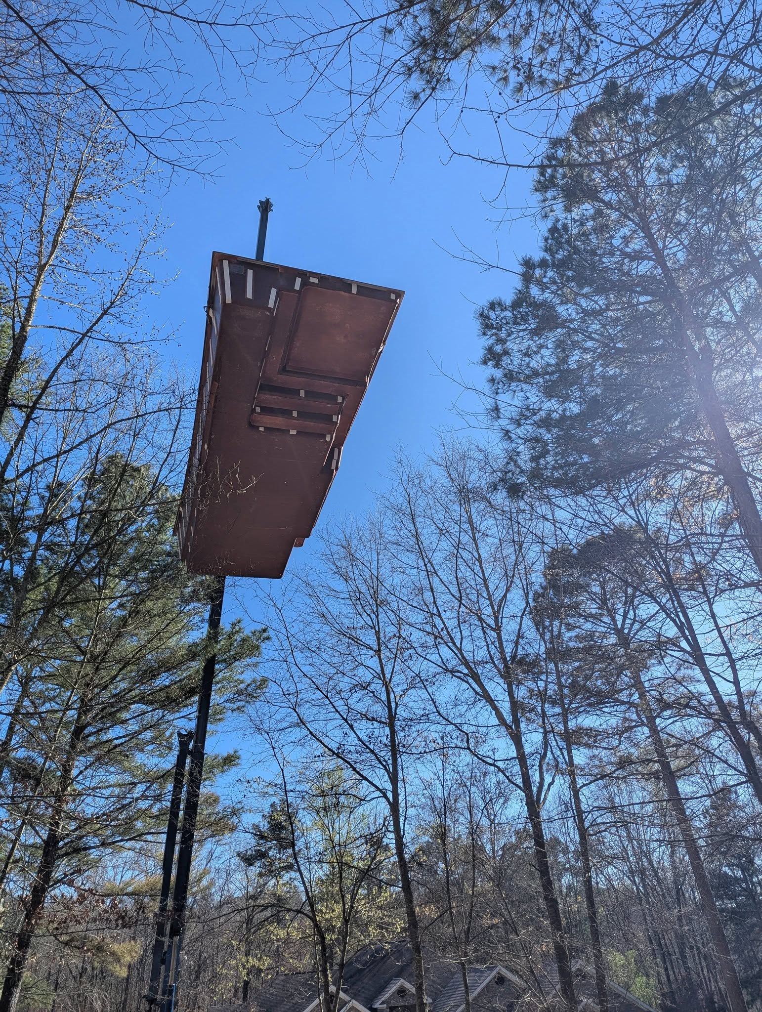 Suspended wooden platform among tall trees under a clear blue sky, with sunlight glare on the right.