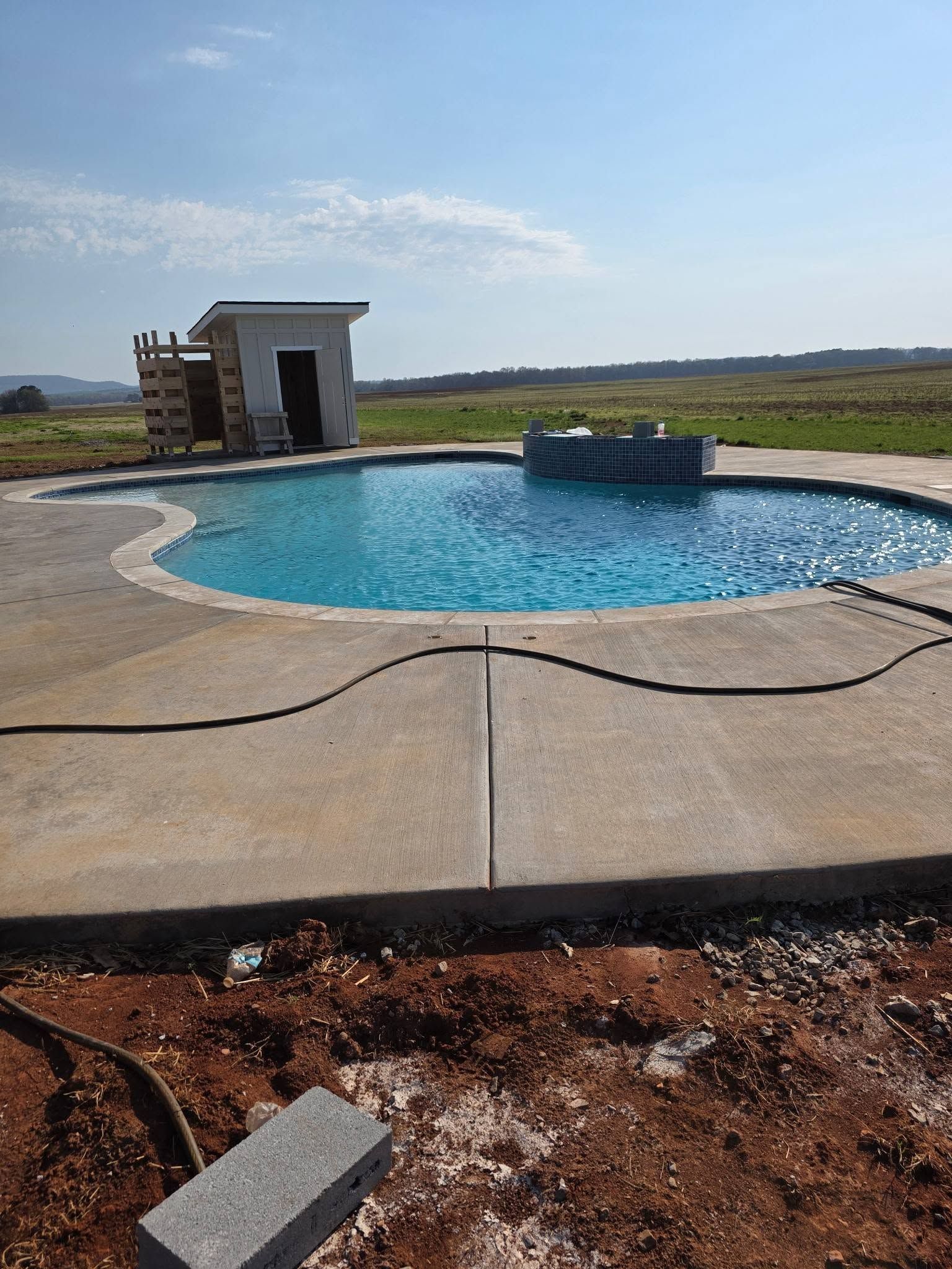 Backyard pool with curved concrete deck, small pool house, and open grassy field beyond