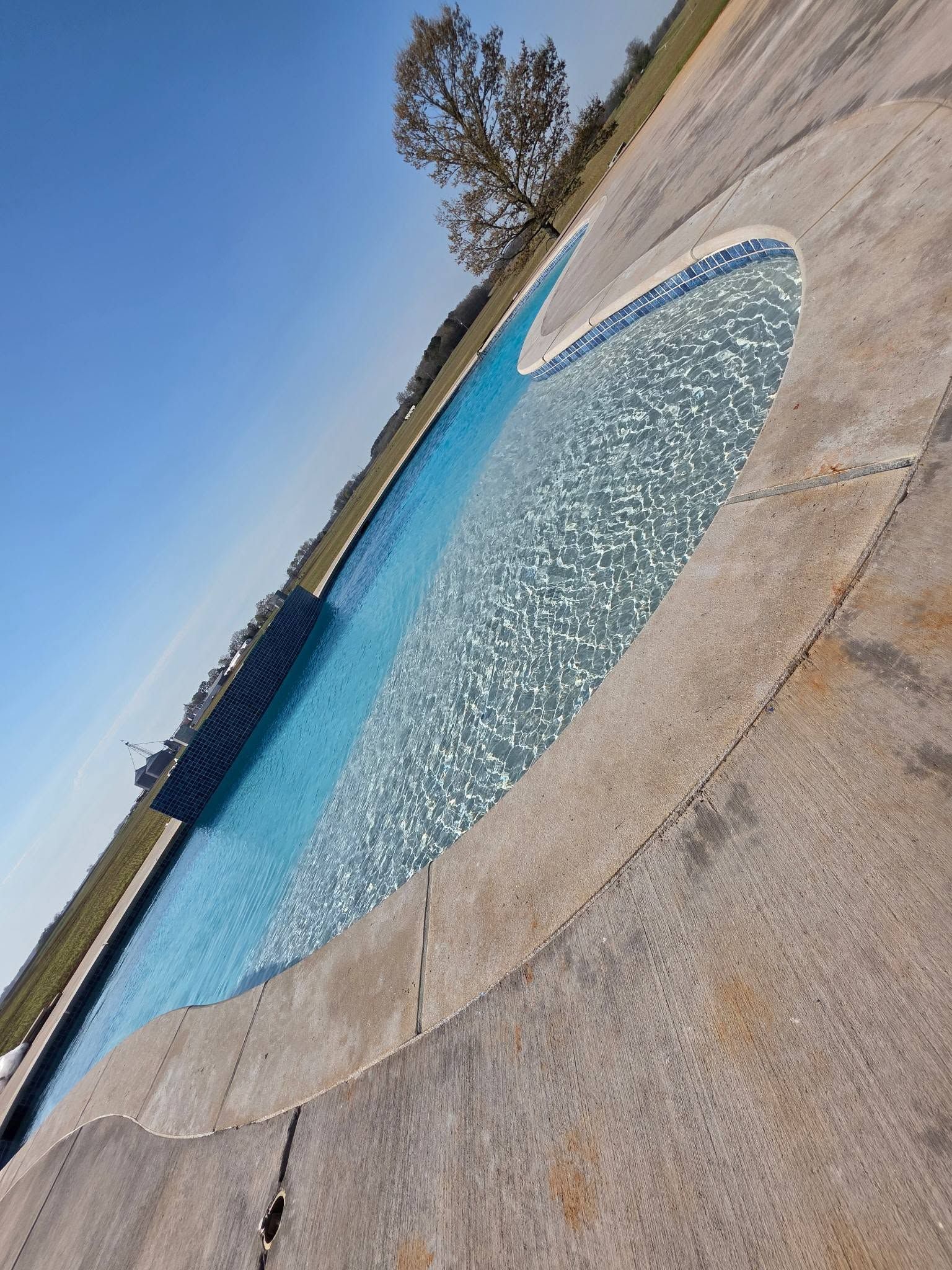 Outdoor swimming pool with blue water and a curved stone deck under a clear sky.
