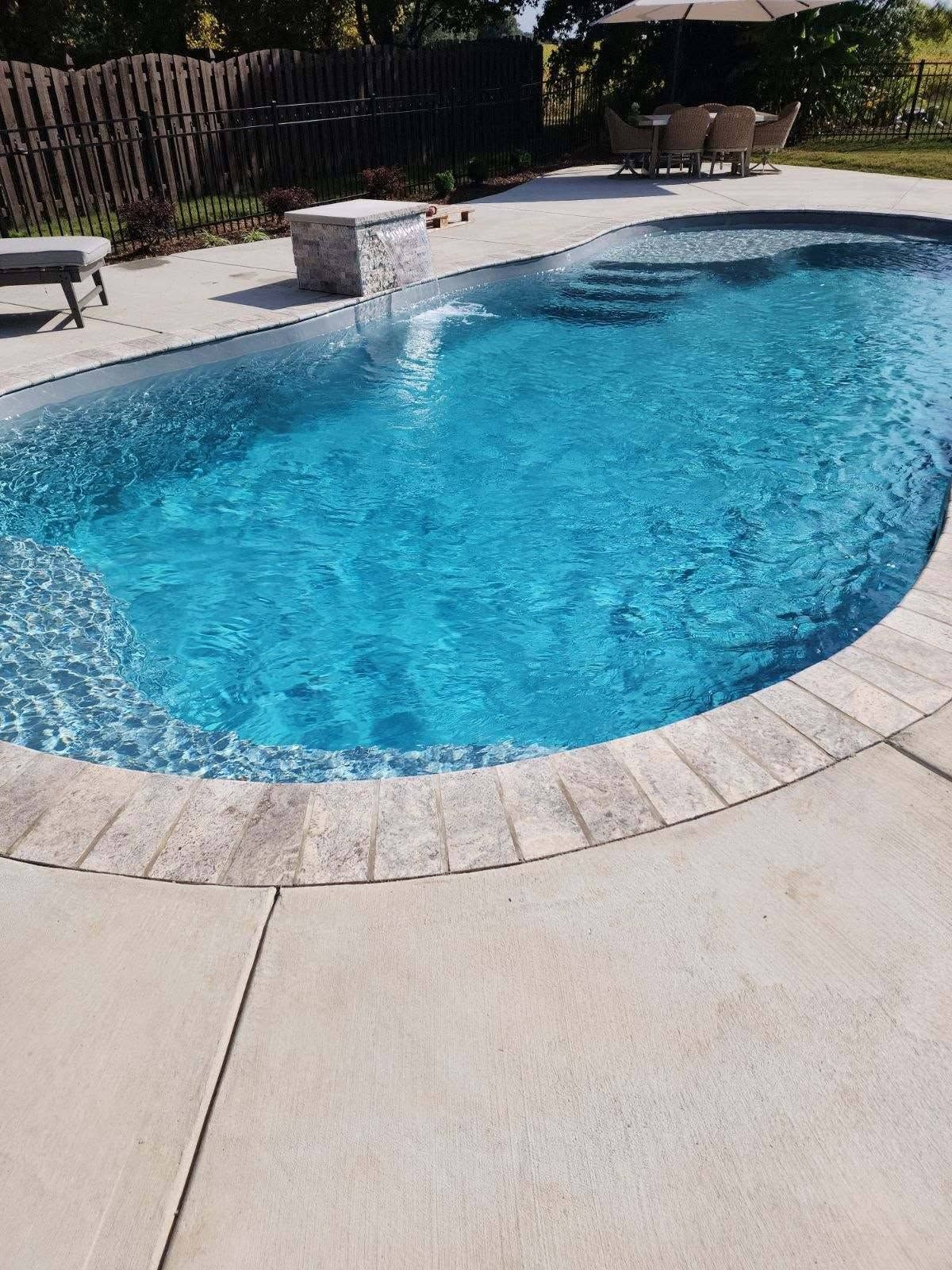 Curved backyard swimming pool with clear blue water and a light stone deck surrounded by a fence