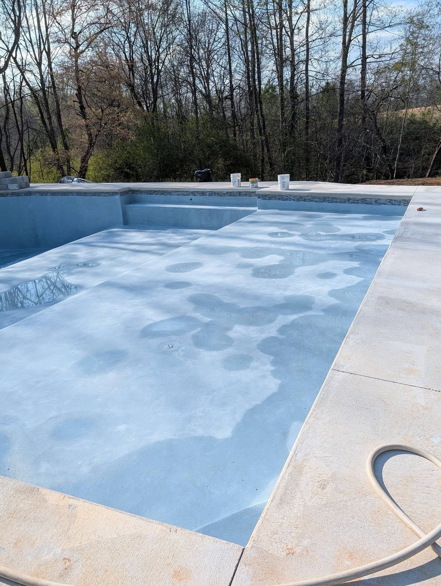 Empty drained swimming pool with blue interior and leaf debris, outdoors by trees.