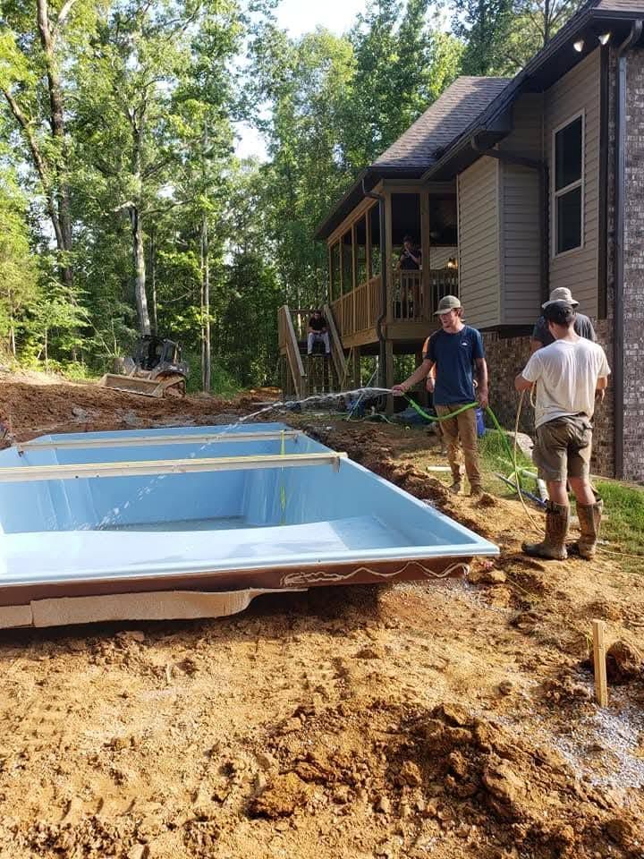Two workers standing beside an in-ground pool shell beside a house in a wooded yard