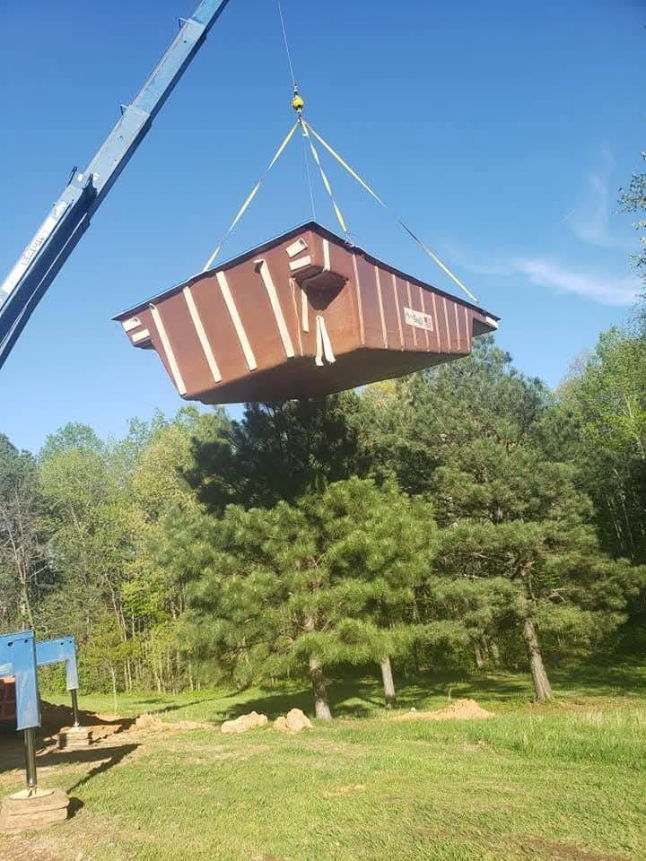 Crane lifting a small brown cabin above a grassy clearing with trees and blue sky.