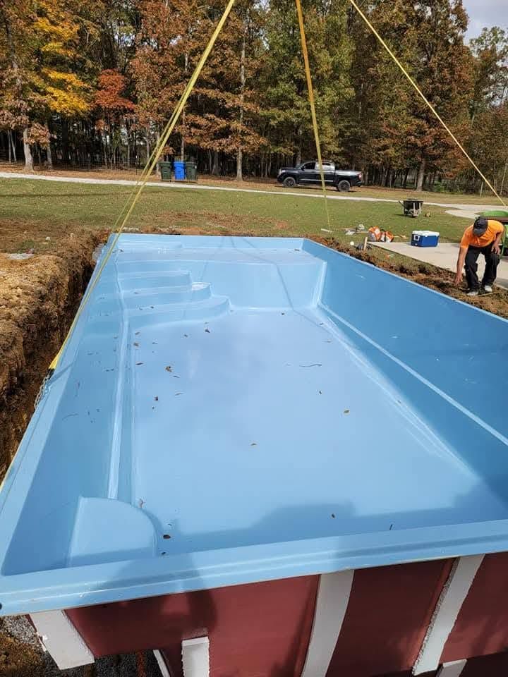 In-ground pool under construction with blue liner, workers nearby, and a wooded yard in fall.