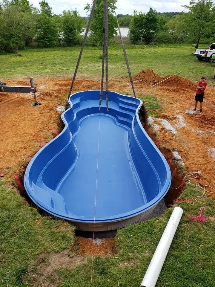Blue fiberglass pool shell being lowered into a dug backyard hole with straps and workers nearby