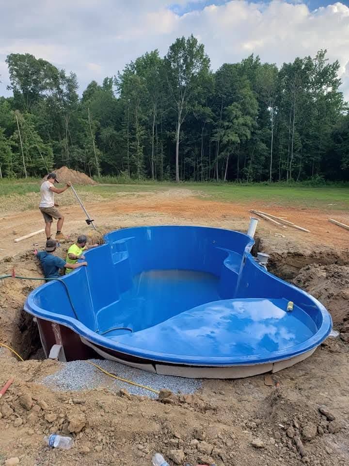 Workers installing a blue in-ground pool in a dirt excavation beside a wooded field
