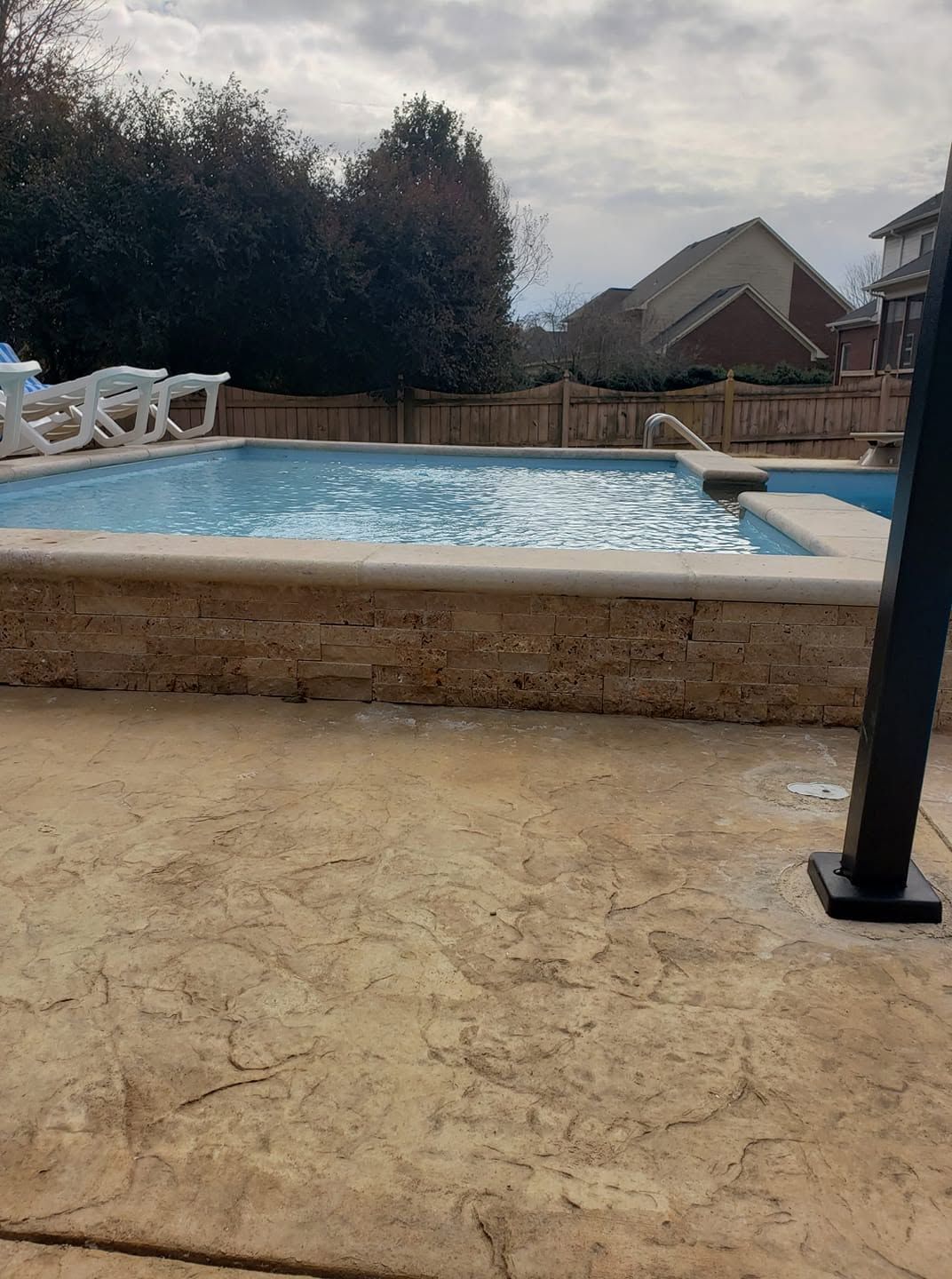 Backyard pool with cloudy water, stone patio, fence, and houses under an overcast sky