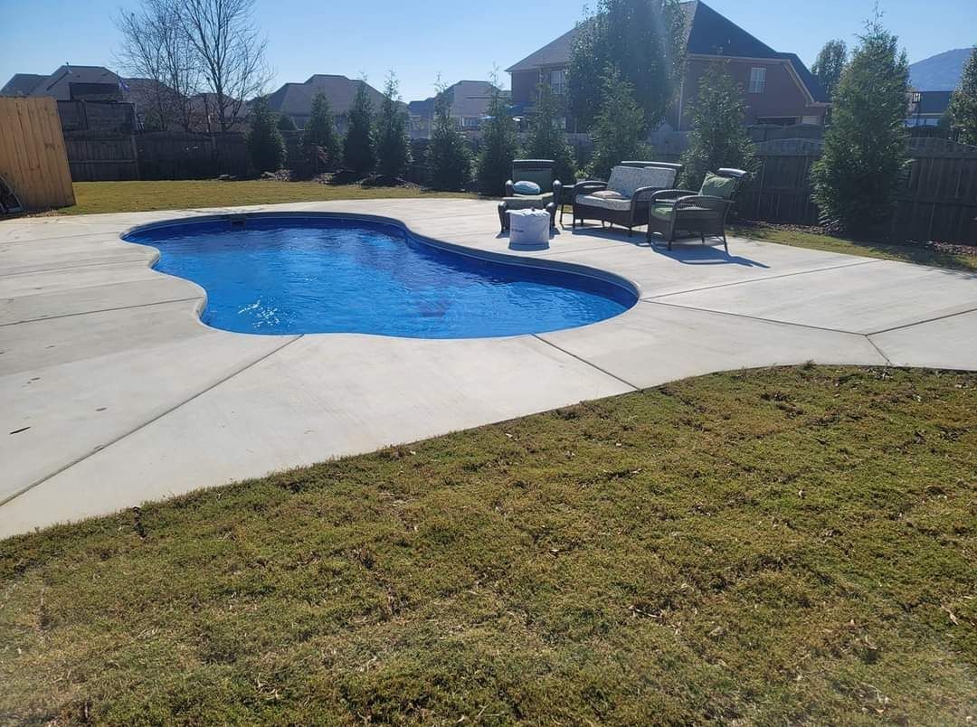 Backyard swimming pool with blue water, concrete patio, lounge chairs, and a grassy lawn in sunlight