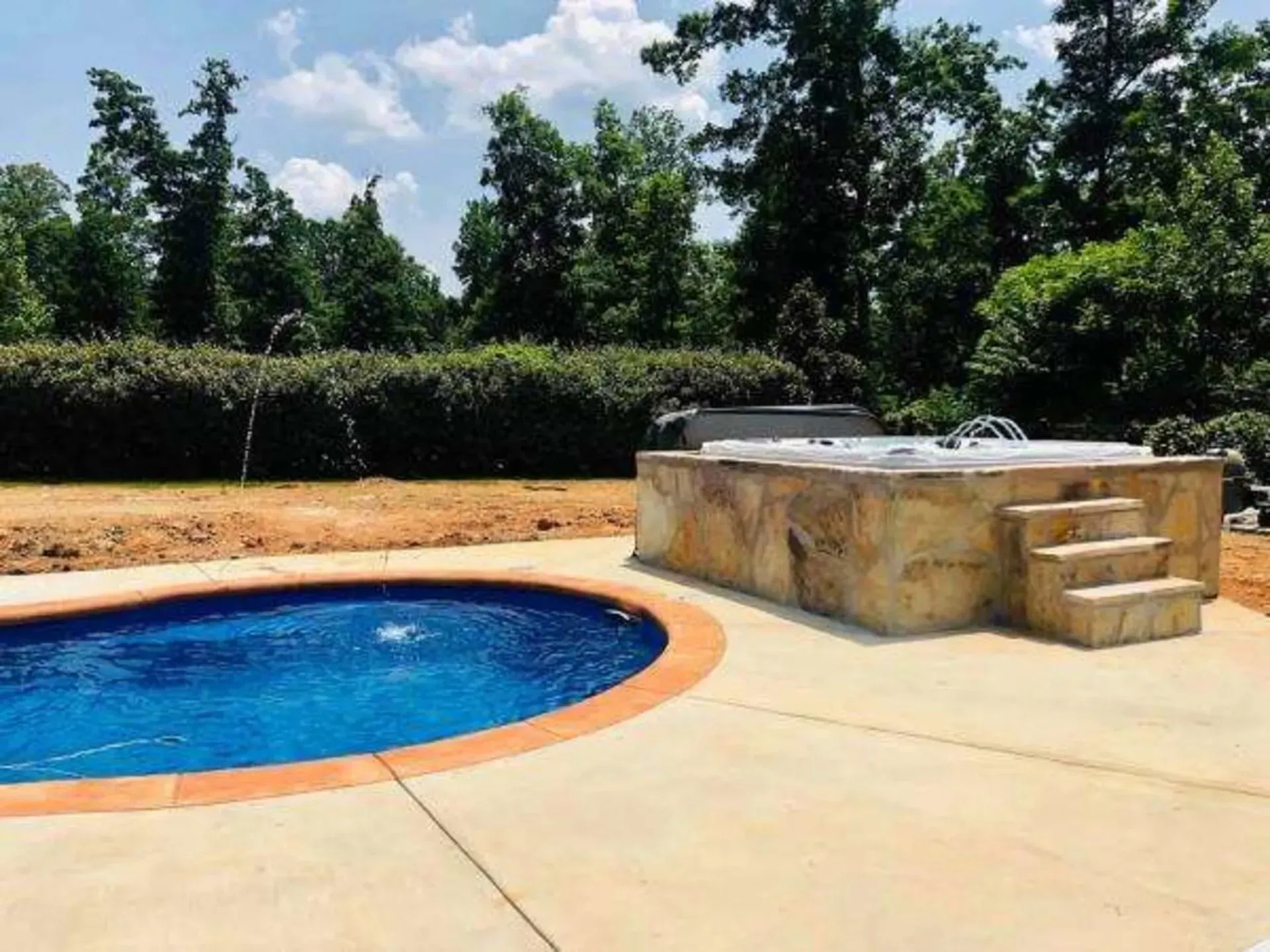 Backyard pool with blue water, tan patio, and raised stone hot tub under trees