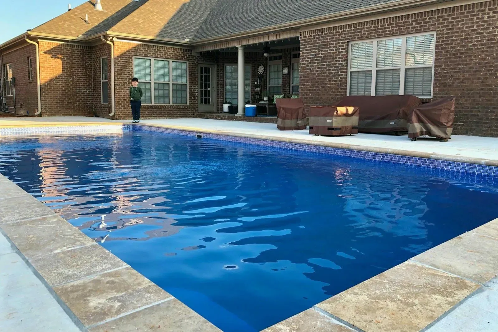 Backyard swimming pool beside a brick house with patio seating and a person near the door