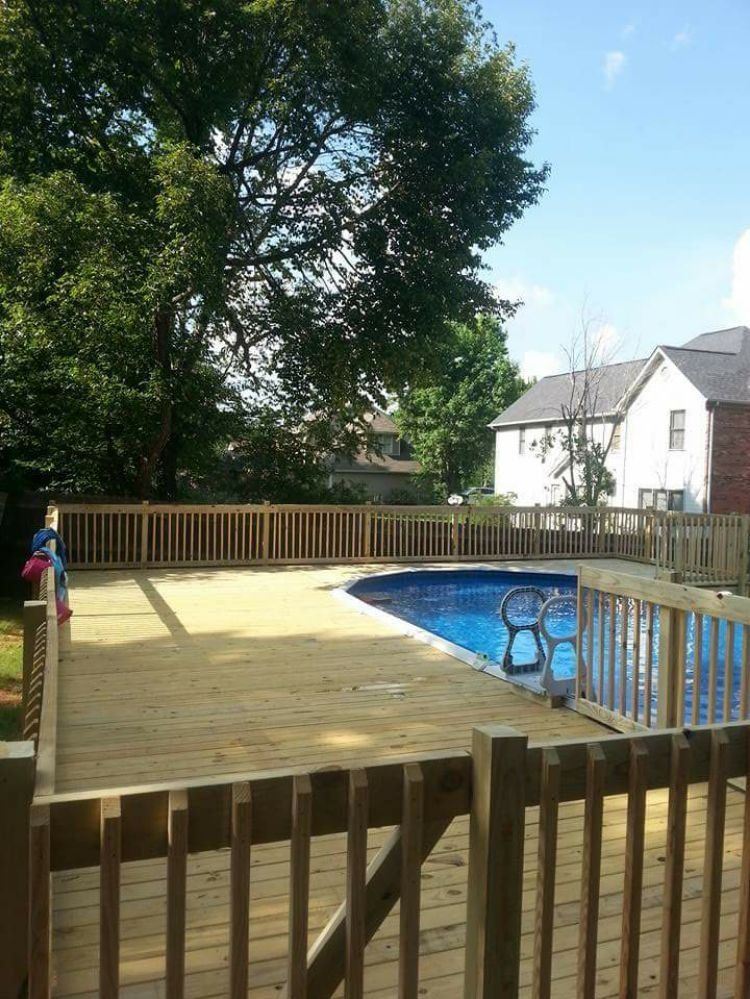 Wooden deck around a blue swimming pool in a backyard, with houses and a large tree nearby.