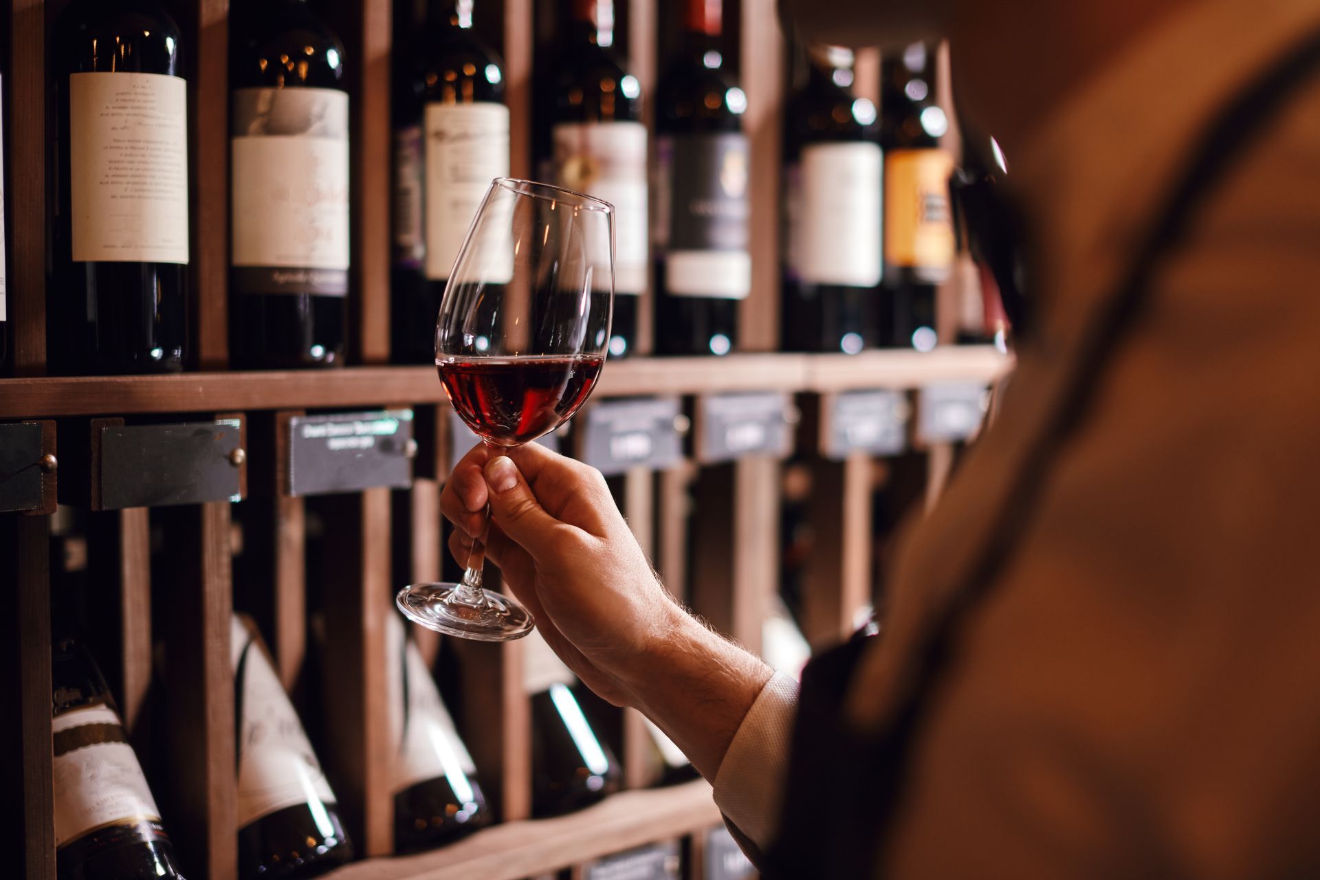 Person holding a wine glass, examining the red liquid, in front of a wine rack.