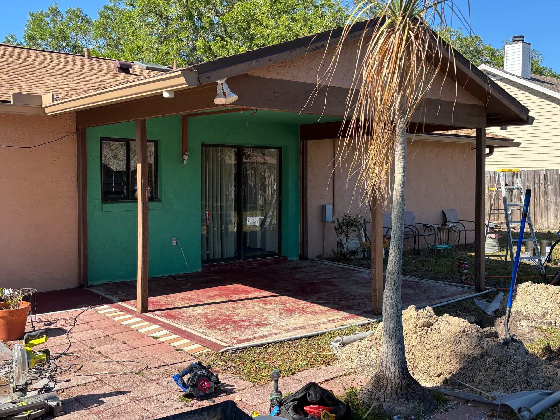 Backyard patio with green doors, covered porch, potted plants, and a small tree in the foreground