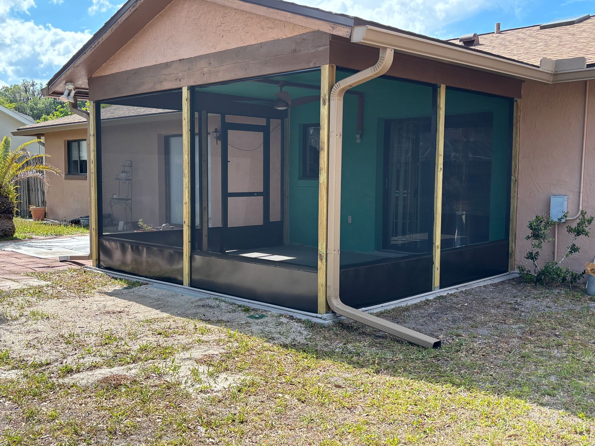 Screened patio enclosure beside a tan house with green trim and gravel yard