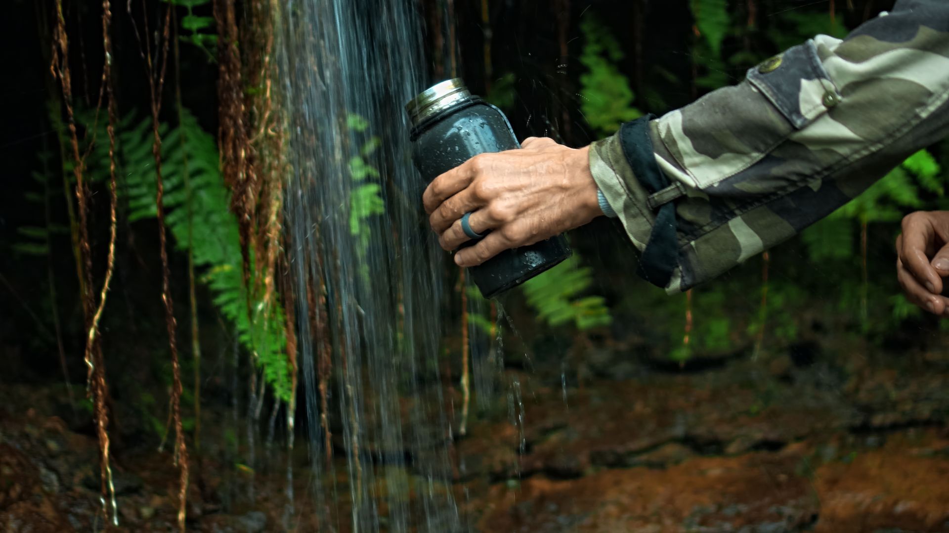 Person filling a dark water bottle from a waterfall in a lush, green forest. They wear camouflage. Person filling a dark water bottle from a waterfall in a lush, green forest. They wear camouflage.
