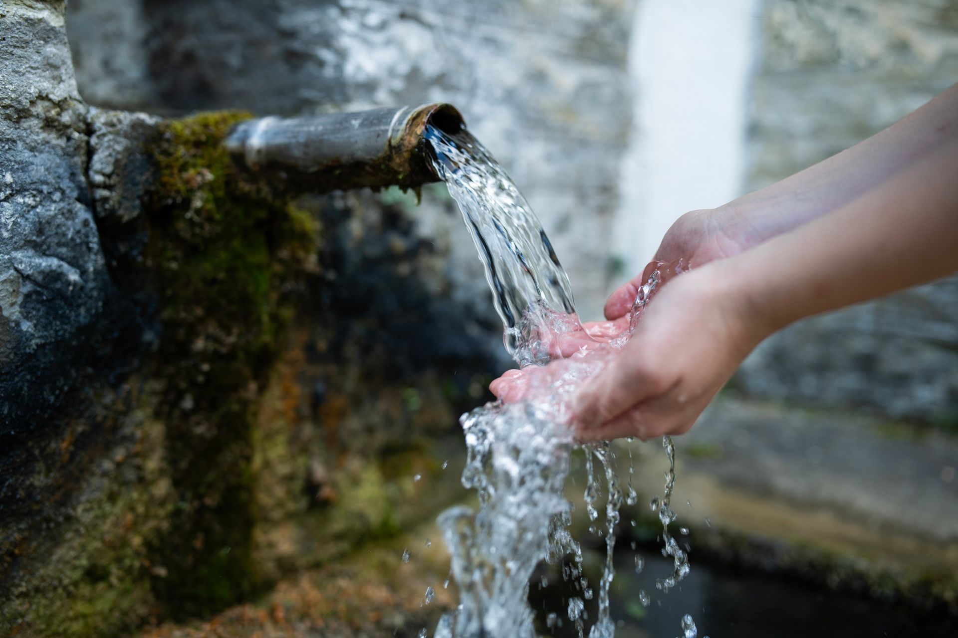 Hands cupped under a metal pipe, catching clear water flowing from a stone fountain. Hands cupped under a metal pipe, catching clear water flowing from a stone fountain.