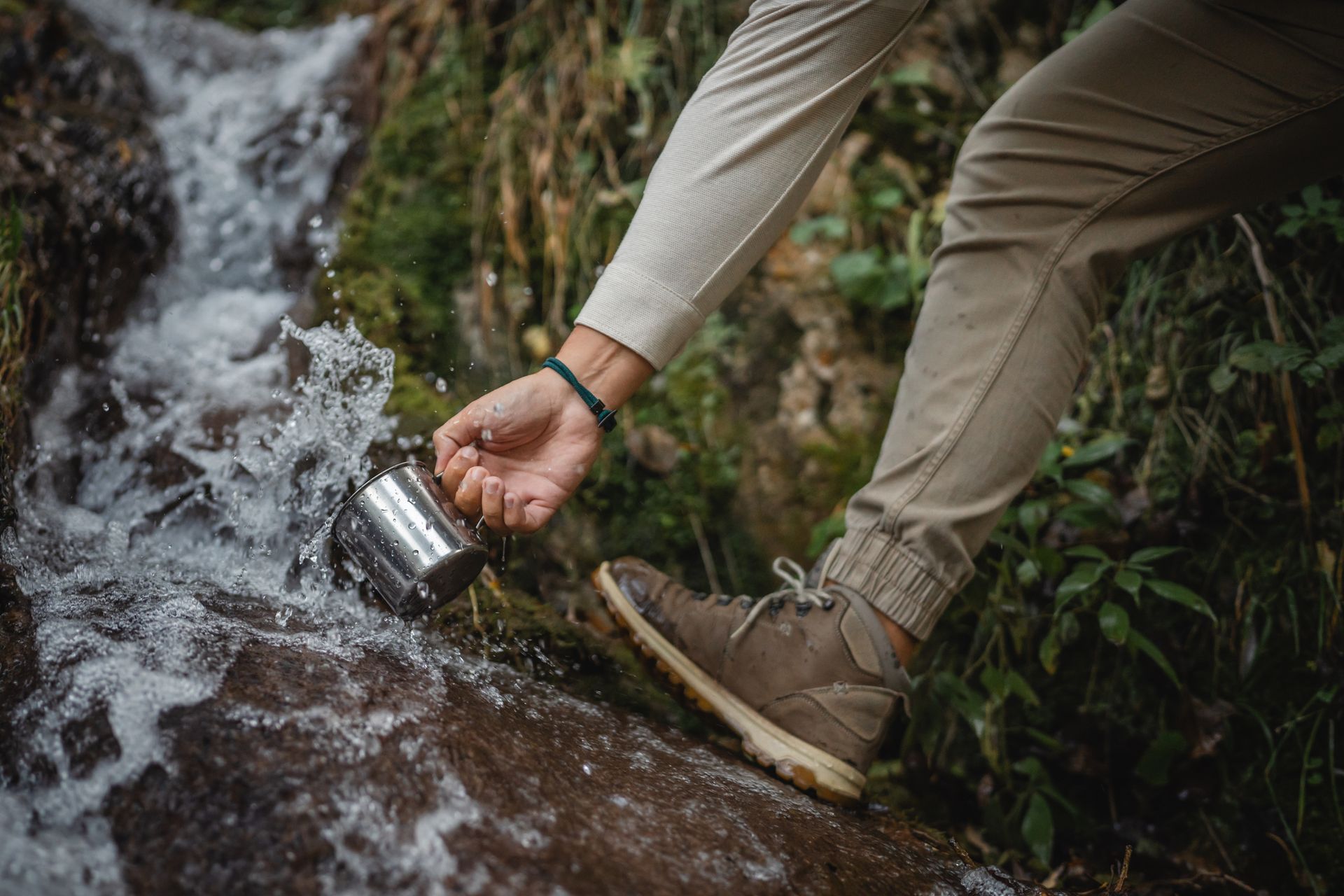 Person scoops water from a flowing stream with a metal cup. They wear tan pants and brown hiking boots in a natural setting. Person scoops water from a flowing stream with a metal cup. They wear tan pants and brown hiking boots in a natural setting.