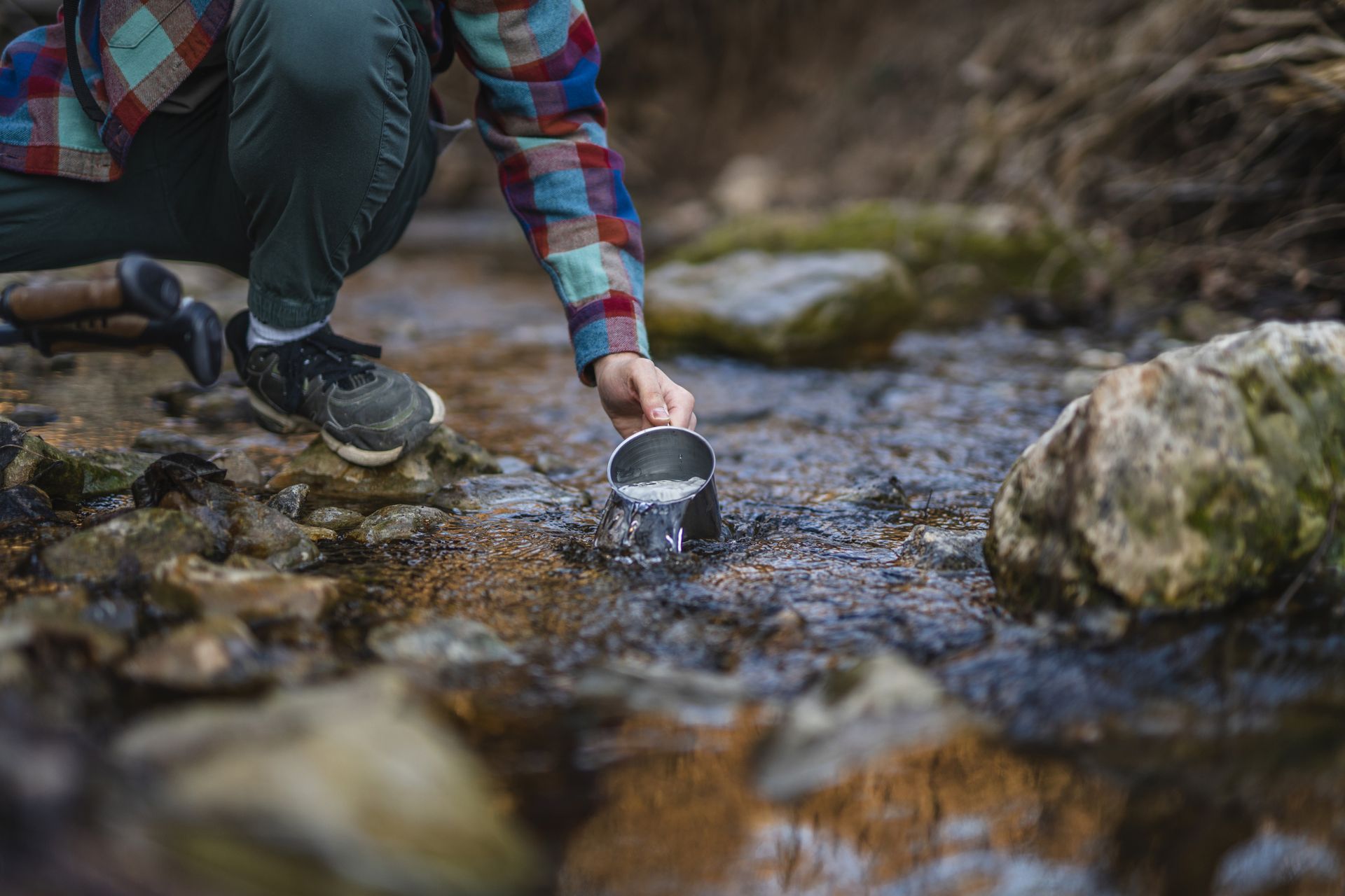 Person kneeling, scooping water from a clear stream with a metal cup in a natural, outdoor setting. Person kneeling, scooping water from a clear stream with a metal cup in a natural, outdoor setting.