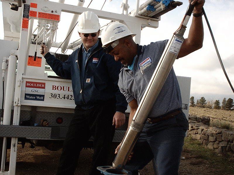 Two men in hard hats and work attire, one holding a long metal tool while working near a truck in an outdoor setting. Two men in hard hats and work attire, one holding a long metal tool while working near a truck in an outdoor setting.
