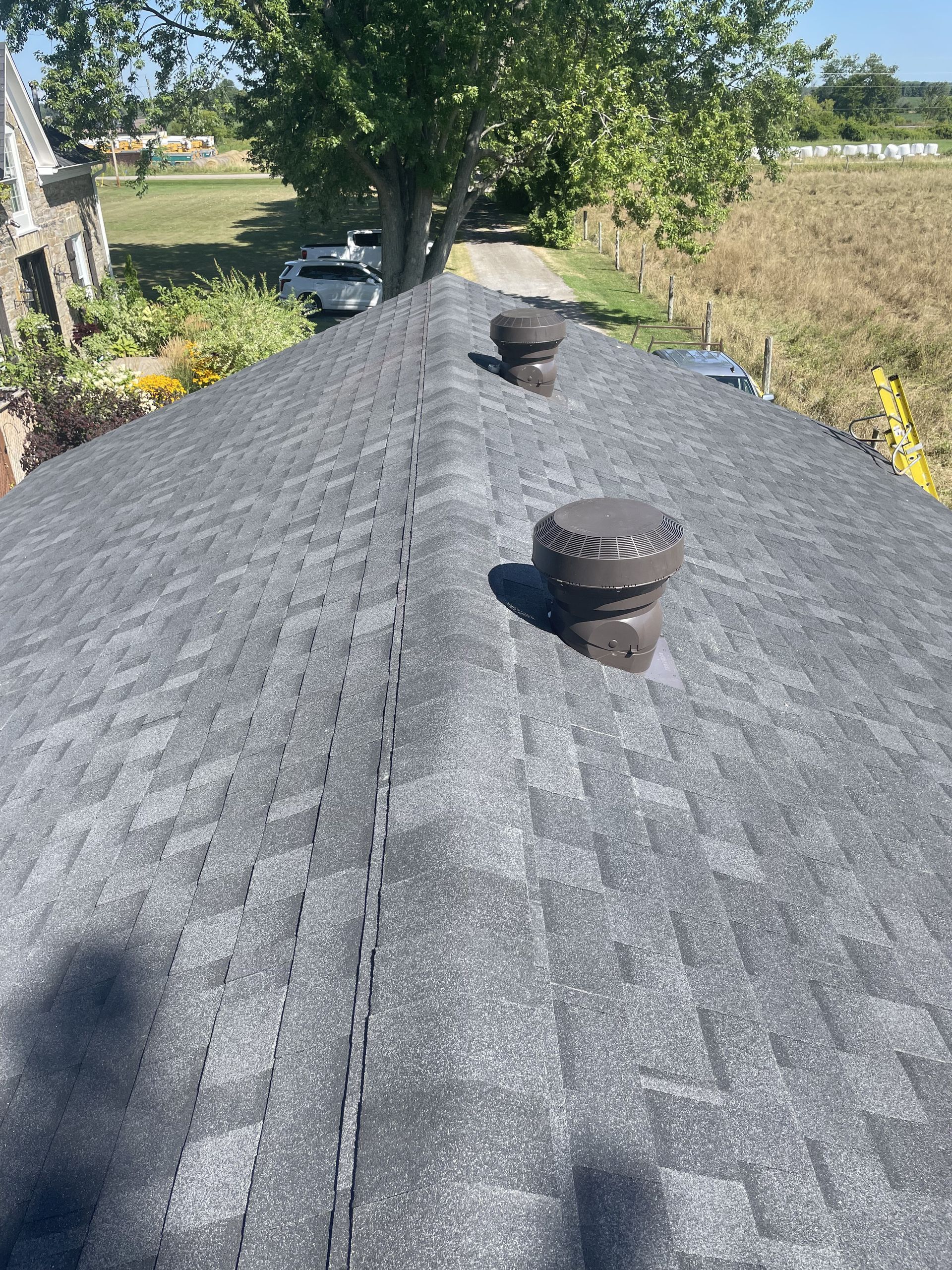 Dark gray shingled roof with two brown vents, a paved driveway, and foliage in the background.