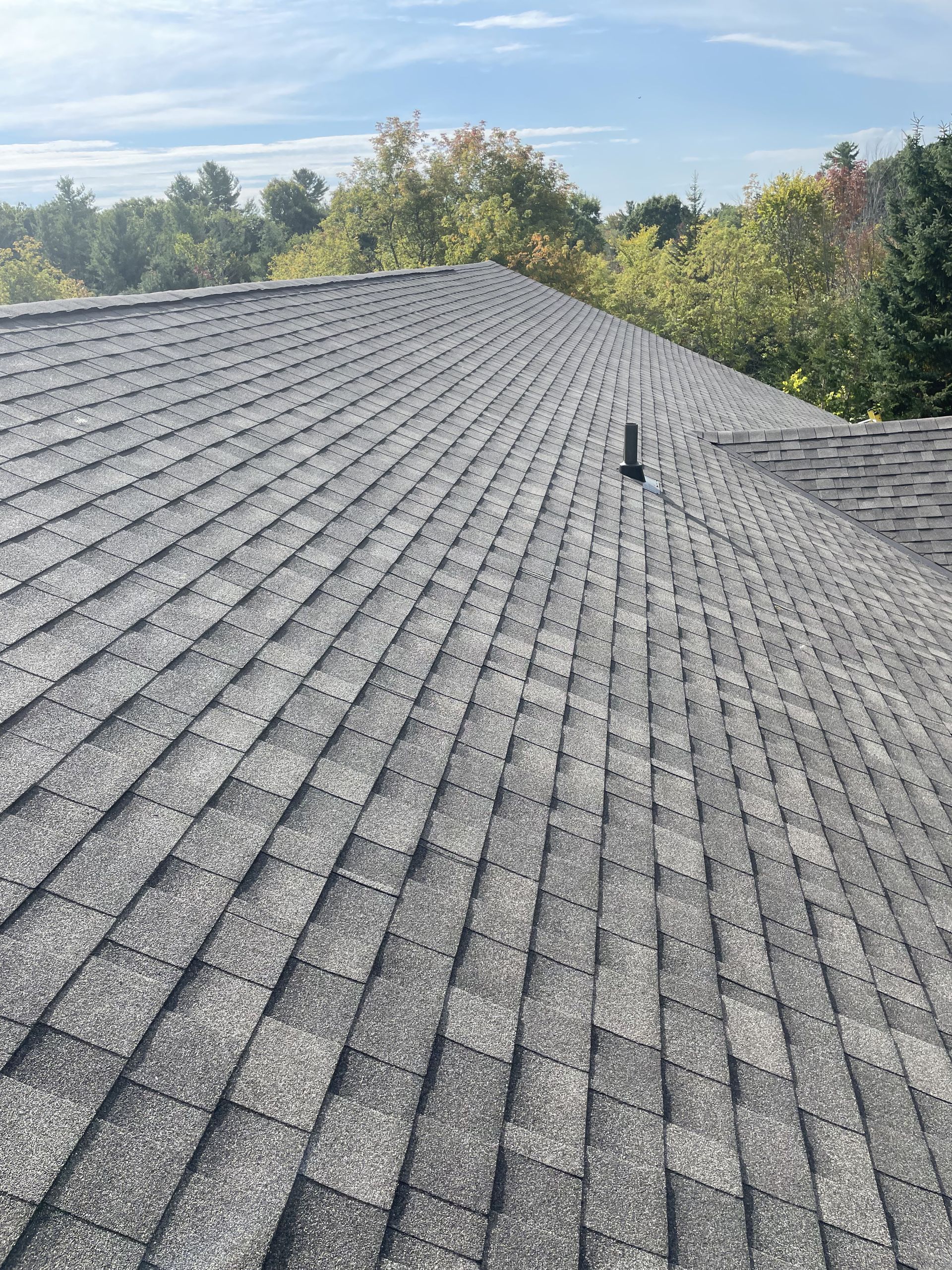 Gray asphalt shingle roof on a house, with a vent pipe and trees in the background.