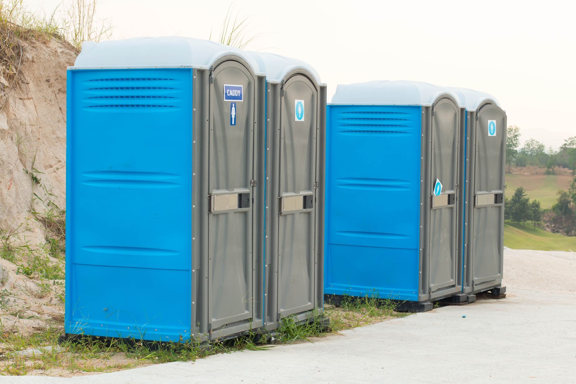Three blue and gray portable toilets lined up outdoors on a paved path.
