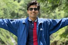 A man wearing sunglasses and a blue jacket is standing with his arms outstretched.