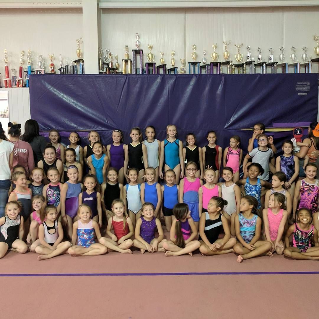 Group of gymnasts posing in rows inside a gym, with trophies displayed along the back wall.