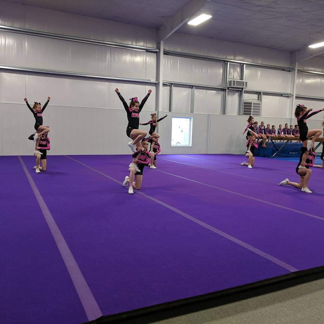 Cheerleaders practicing stunts on a purple gym mat inside an indoor training facility