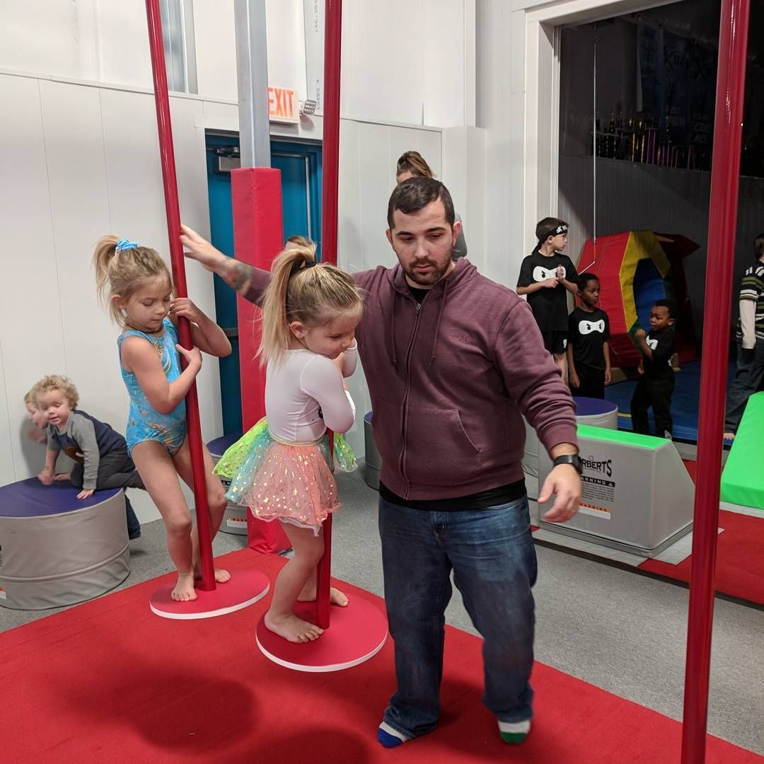 Man assisting two children on circus-style poles in a red indoor play area