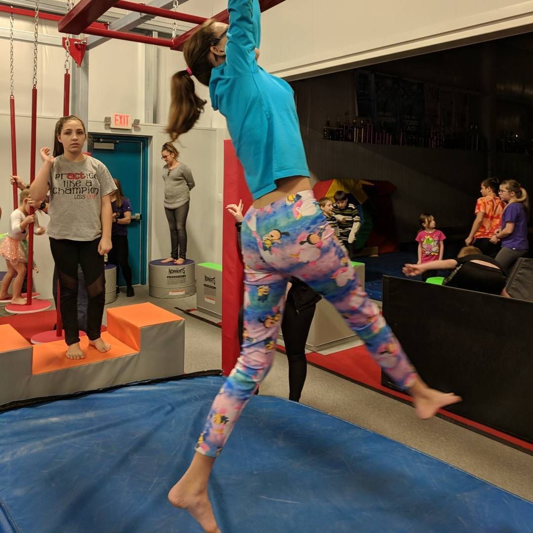 Child swinging on a rope in a gym, balancing on a blue mat while others watch nearby