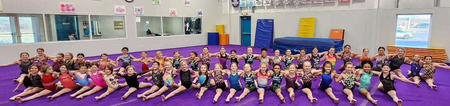 Large group seated on a gym floor in purple outfits, posing together indoors.