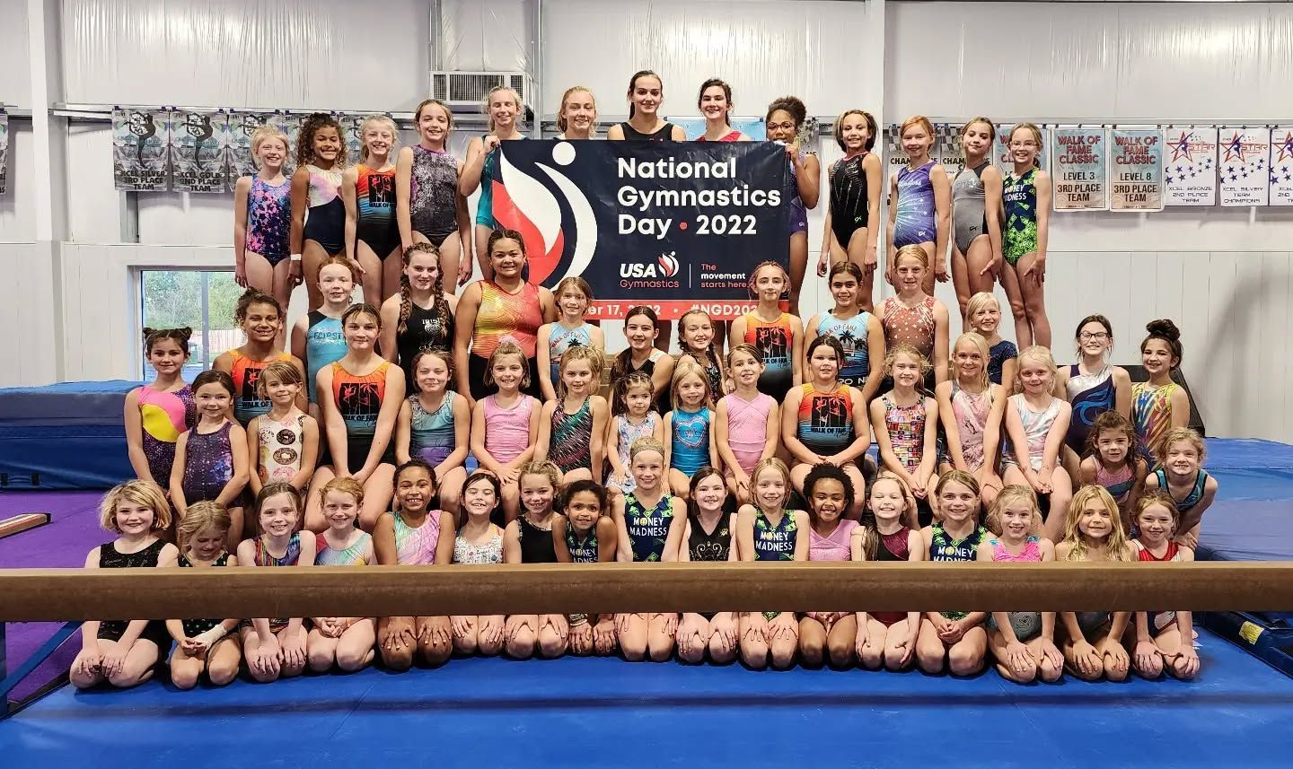 Large gymnastics team group photo in a gym, posing with a banner on a blue mat.