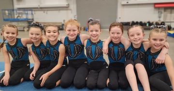 Seven young gymnasts in blue-and-black leotards sitting in a row, smiling in a gym.