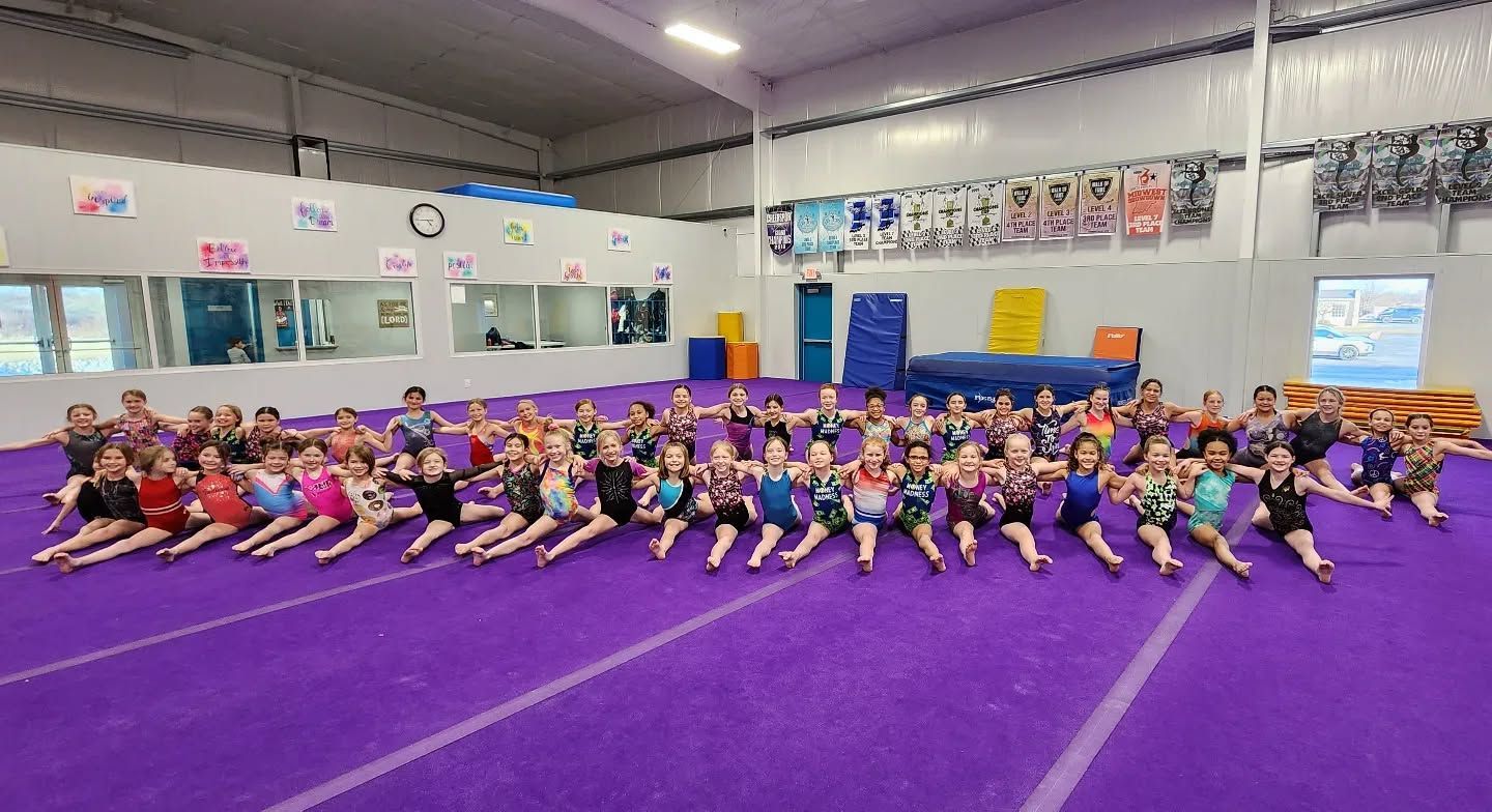 Large group stretching on purple mats in a gym, with a mirror wall and posters in the background