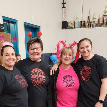 Four people smiling indoors, wearing black and pink shirts with red heart decorations in the background