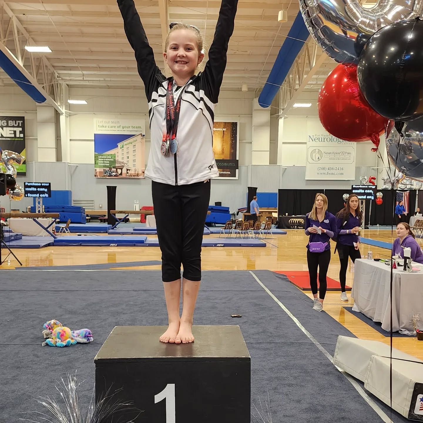 Gymnast on podium with arms raised at an indoor competition, wearing a medal and white jacket.