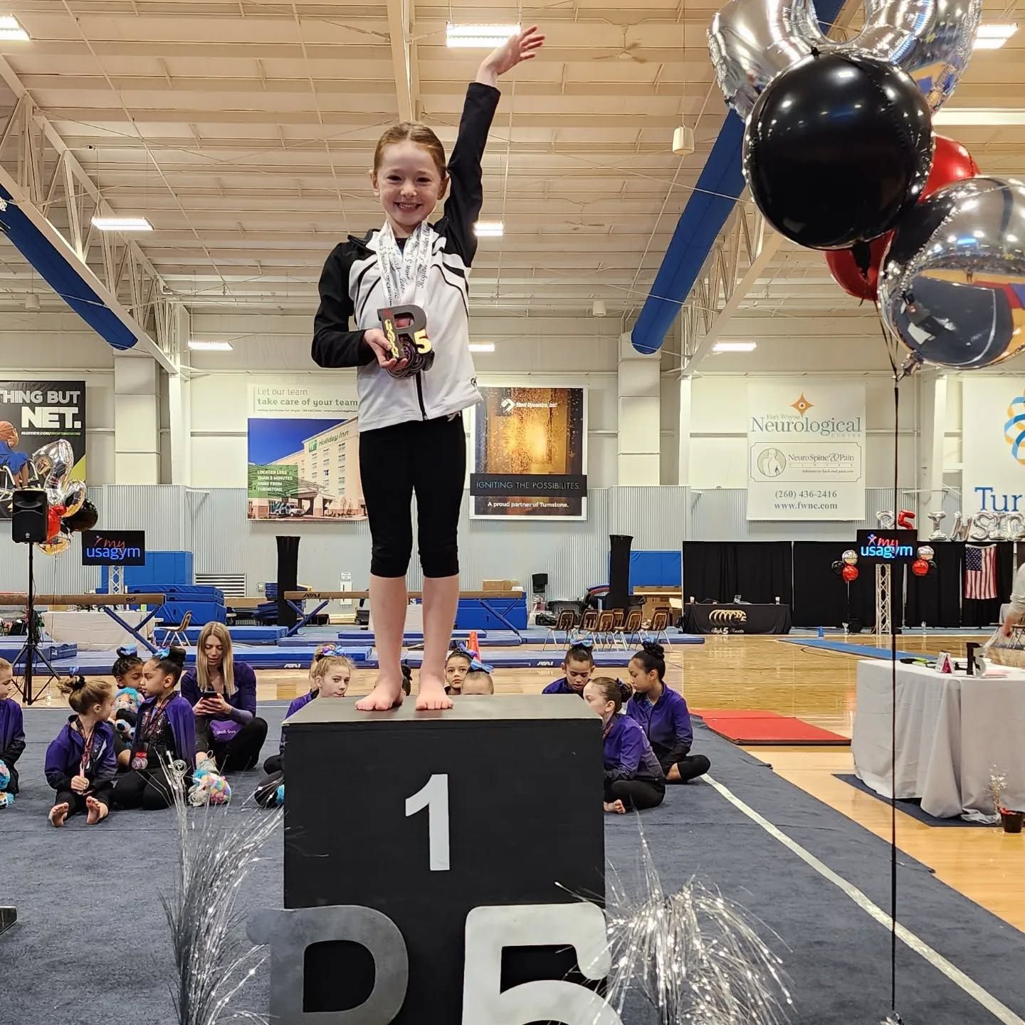 Young gymnast stands on first-place podium with medal and arm raised at a competition inside a gymnasium.