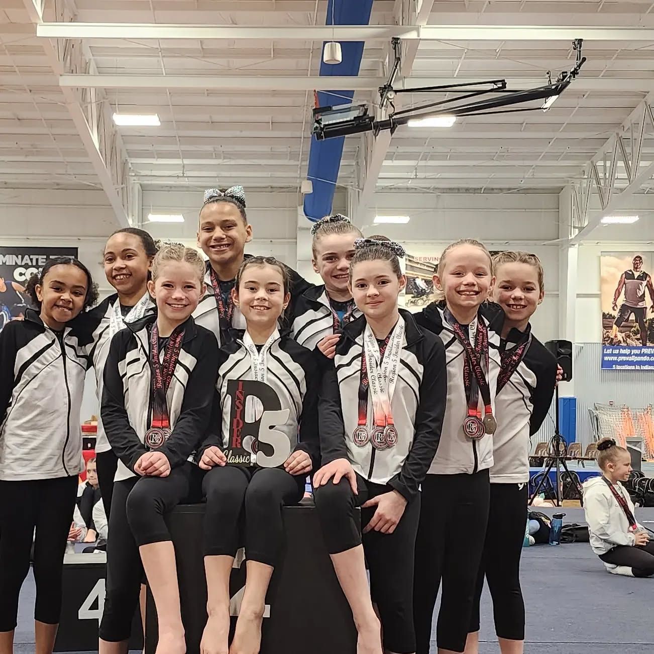 Group of young gymnasts posing in a gym, wearing black-and-white team uniforms and medals.