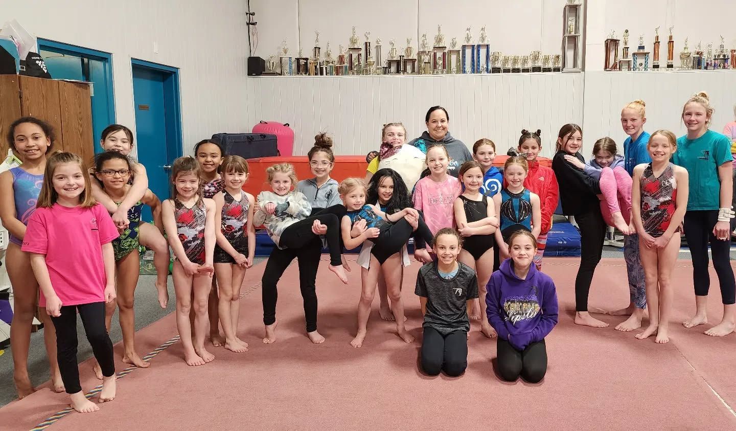 Group of children and adults posing in a dance studio with trophies on shelves behind them.