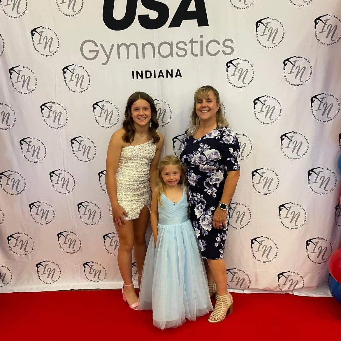 Three people posing on a red carpet at a USA Gymnastics Indiana event backdrop.