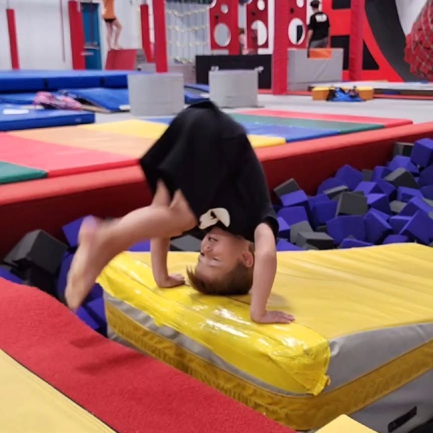 Child doing a headstand on a yellow mat in a colorful indoor gym with foam pits and trampolines