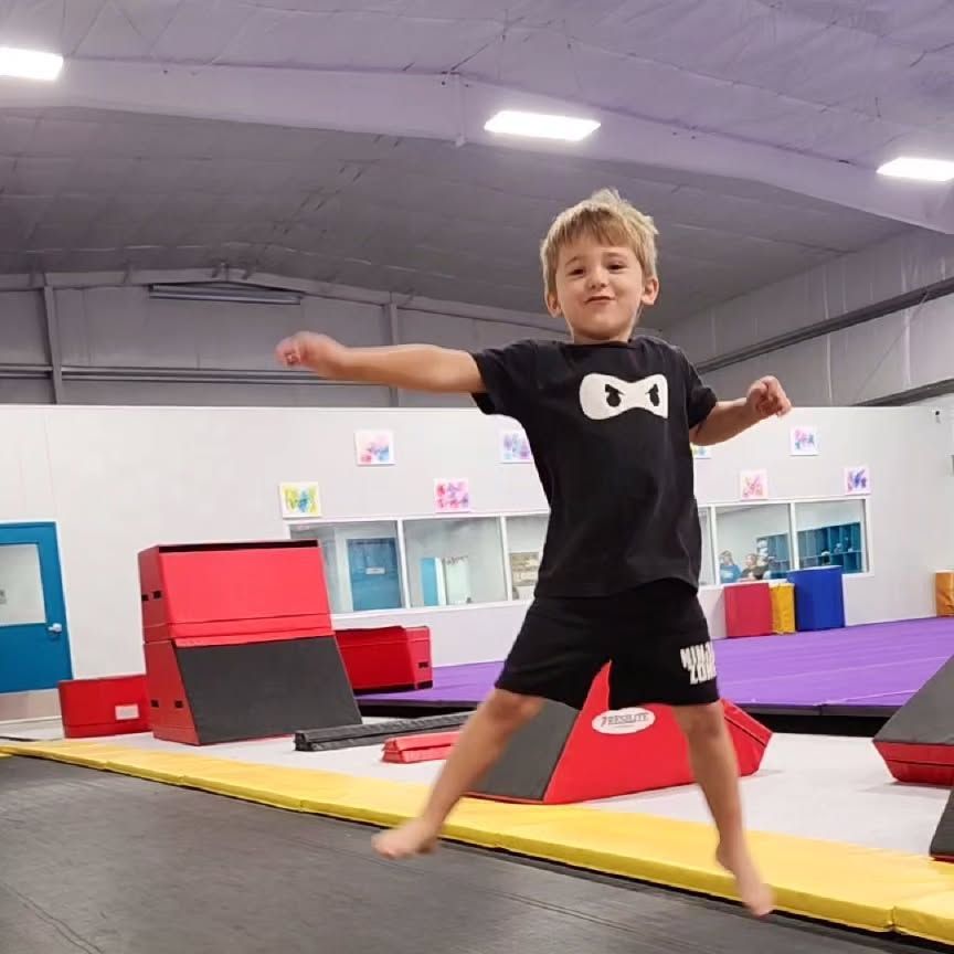 Child balancing on a yellow padded beam in an indoor gymnastics gym, arms outstretched, with red and purple mats nearby