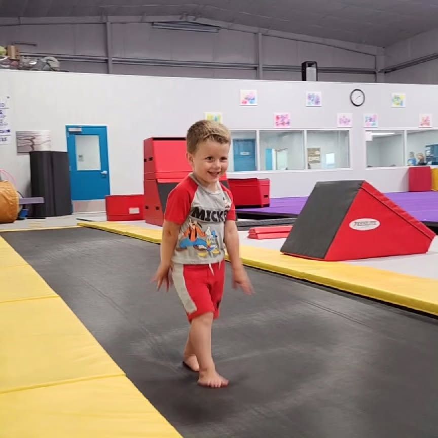 Child smiling on a trampoline in a gym, with red and yellow padded mats nearby.