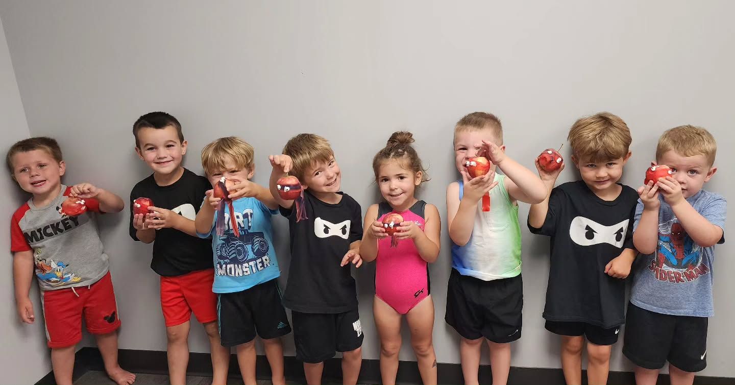 Eight children standing in a row, holding colorful drinks and posing against a plain wall.