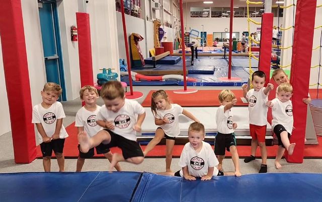 Children posing in a gym with red and blue padding, some crouching and smiling toward the camera