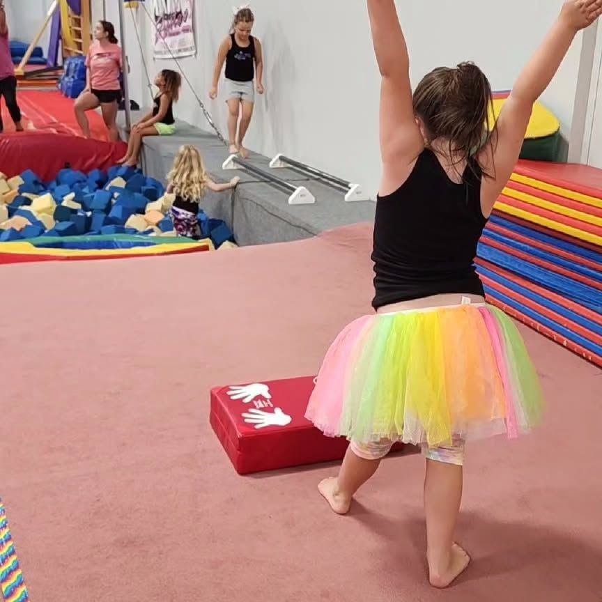 Child in a rainbow tutu stands with arms raised in a gymnastics play area.