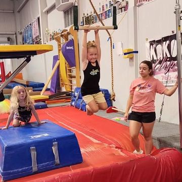 Children in a gymnastics gym on mats and uneven bars, one hanging from rings, others watching nearby