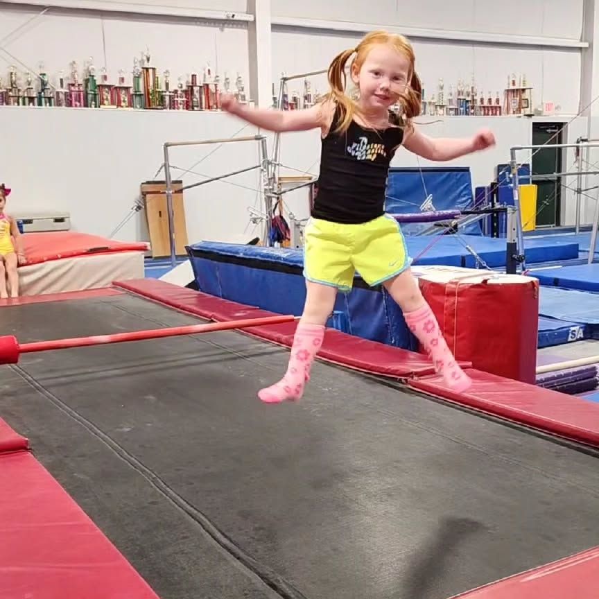 Child mid-jump on an indoor gymnastics trampoline, wearing a black top and neon yellow shorts