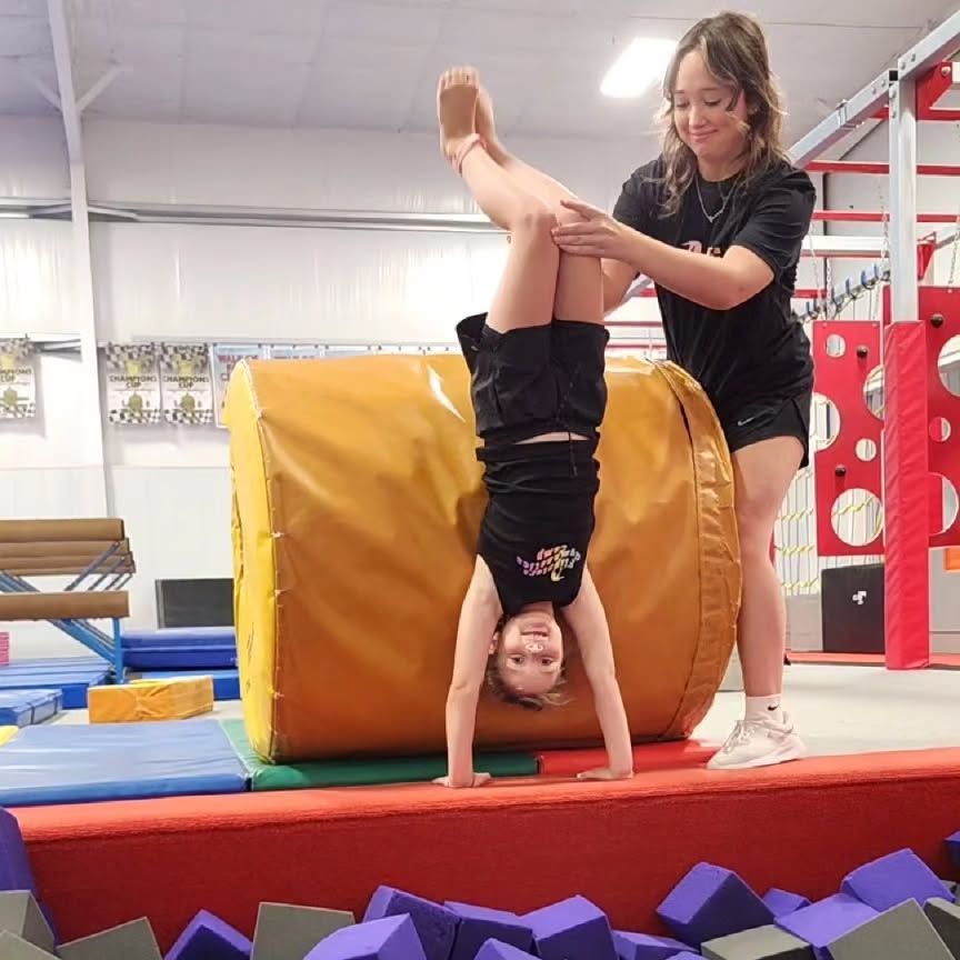 Gymnast helping a child balance upside down on a padded cylinder in an indoor gym